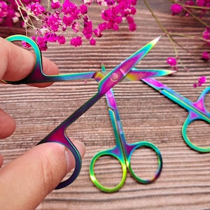 May include: Three rainbow-colored stainless steel cuticle scissors with sharp blades. The scissors are on a wooden surface.