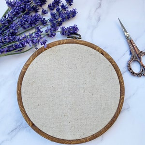 Pu&ograve; includere: Un cerchio da ricamo in legno marrone con un tessuto di lino beige teso al centro. Il cerchio &egrave; su una superficie di marmo bianco con fiori di lavanda e un paio di forbici ornate.