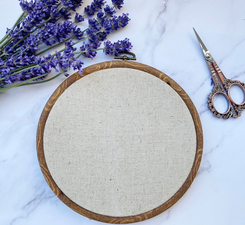Pu&ograve; includere: Un cerchio da ricamo in legno marrone con un tessuto di lino beige teso al centro. Il cerchio &egrave; su una superficie di marmo bianco con fiori di lavanda e un paio di forbici ornate.