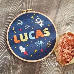 May include: A navy blue embroidered hoop art piece with the name "LUCAS" in colourful thread. The design features an astronaut, planets, stars, and a rocket ship. The hoop is set in a wooden frame.