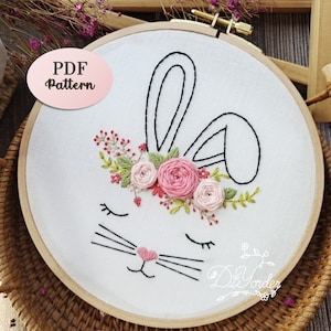 May include: A white embroidery hoop featuring a black line drawing of a bunny with a floral crown. The bunny has closed eyes and a pink heart nose. The floral crown is made of pink and white flowers and green leaves.