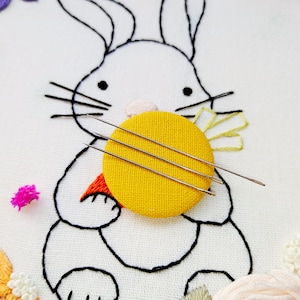 May include: Embroidered artwork featuring a white bunny with black outlines, holding a yellow circle with three needles. The bunny has a carrot and flower details. The background is white fabric with additional floral embroidery.