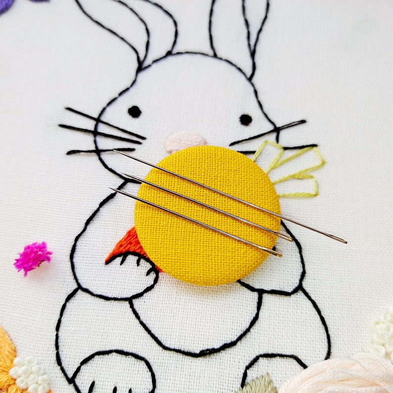May include: Embroidered artwork featuring a white bunny with black outlines, holding a yellow circle with three needles. The bunny has a carrot and flower details. The background is white fabric with additional floral embroidery.