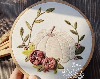 May include: Embroidered hoop art featuring a beige pumpkin with green leaves and pink flowers. The hoop is made of wood and has a white fabric background.