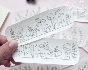 Op de afbeelding: Een set witte papieren vellen met zwarte lijntekeningen van paddenstoelen en bladeren. De vellen zijn getextureerd en hebben een subtiel patroon.