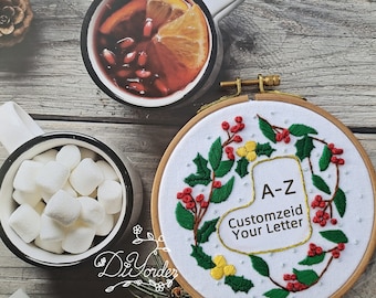 May include: Embroidered hoop art with a white background, featuring a green holly sprig with red berries and yellow flowers. The hoop is made of wood and has a gold metal ring. The text "A-Z Customize Your Letter" is embroidered in the center of the hoop.