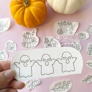 Op de afbeelding: Een wit stoffen vel met drie getekende geesten met open monden en boze ogen. Het vel wordt vastgehouden door een hand. Het vel is omringd door andere witte stoffen vellen met verschillende herfstthema's.