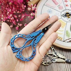 Pode incluir: Tesoura de bordado de metal azul com um design floral decorativo. As tesouras têm uma ponta pontiaguda e são seguradas na mão.
