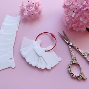 Op de afbeelding: Witte plastic draadklossen met een rode elastische lus en een paar zilveren decoratieve scharen op een roze achtergrond.