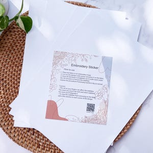 May include: A stack of white embroidery stabiliser sheets with an instructional card titled Embroidery Sticker. The card features floral line art, a QR code, and text detailing how to use the stabiliser for fabric projects and stitching.