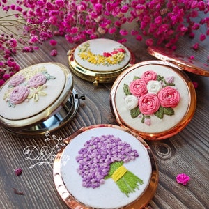 May include: Four embroidered compact mirrors with floral designs. The mirrors have gold and rose gold trim. The floral designs include a wreath of yellow flowers, a bouquet of pink roses, a bouquet of purple flowers, and a bouquet of white and pink roses.