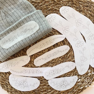 May include: A gray knit beanie with a floral design patch, surrounded by several white fabric patches with floral embroidery. The patches vary in shape and size, with some curved and others rectangular. The items are arranged on a woven, brown surface.
