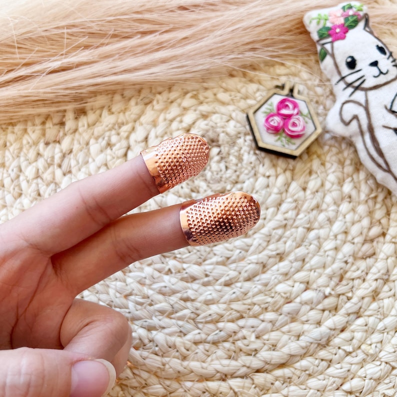 May include: Two copper-colored thimbles with a textured surface are worn on two fingers. The thimbles are displayed against a woven background, with a small embroidered piece and a cat-shaped plush toy in the background.