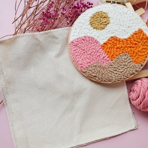 May include: A white canvas with a brown wooden embroidery hoop with a colorful landscape design. The design features a yellow sun, pink and orange mountains, and brown hills. The hoop is surrounded by dried flowers and pink yarn.