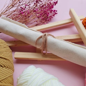May include: A rolled up piece of white canvas fabric tied with a brown string, sitting on a wooden frame. There are two balls of yarn, one brown and one white, in the foreground.