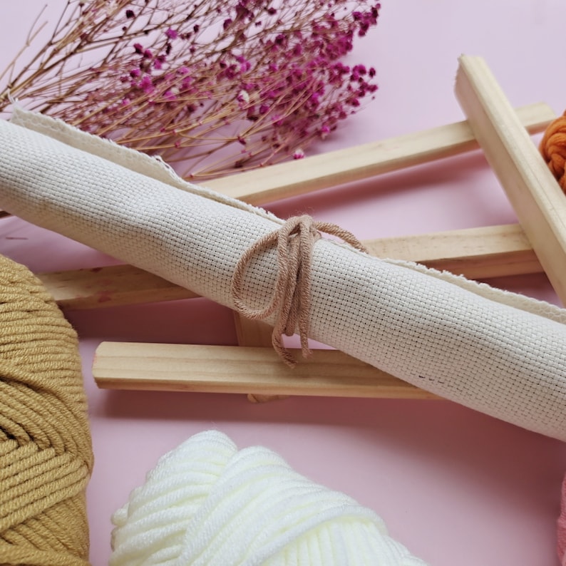 May include: A rolled up piece of white canvas fabric tied with a brown string, sitting on a wooden frame. There are two balls of yarn, one brown and one white, in the foreground.
