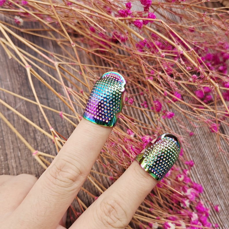 May include: Two iridescent metal thimbles, each worn on a finger. The thimbles are covered in small, evenly spaced holes. The colors shift from blue to green to purple. The background is a wooden surface with dried flowers.
