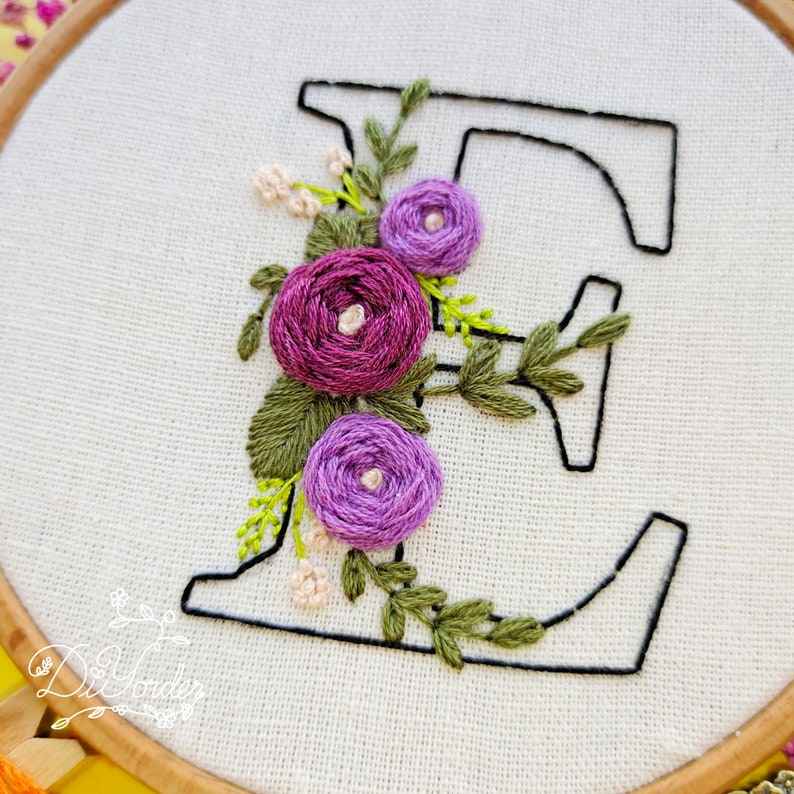 May include: Embroidered letter E with purple and white flowers and green leaves. The letter is stitched on a white fabric background.