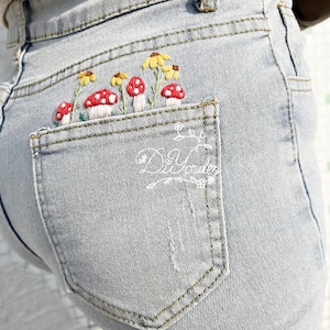 Peut inclure: Un jean en denim bleu clair avec une poche arrière brodée de champignons rouges et de fleurs jaunes.