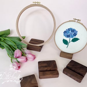 May include: Two embroidery hoops with wooden stands. The smaller hoop features a blue and white embroidered flower design. The larger hoop is empty. Both hoops are on a light pink surface.