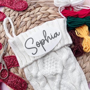 May include: White knitted stocking with a personalised name tag that says "Sophia". The stocking is surrounded by colourful yarn for embroidery.