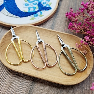 May include: Three pairs of small, decorative scissors with ornate handles. The scissors are gold, copper, and silver in color and are arranged on a wooden tray.