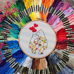Pu&ograve; includere: Un cerchio da ricamo bianco con un disegno di due orsi che tengono cuori. Gli orsi sono circondati da fiori e foglie. Il cerchio &egrave; circondato da una variet&agrave; di fili da ricamo colorati.