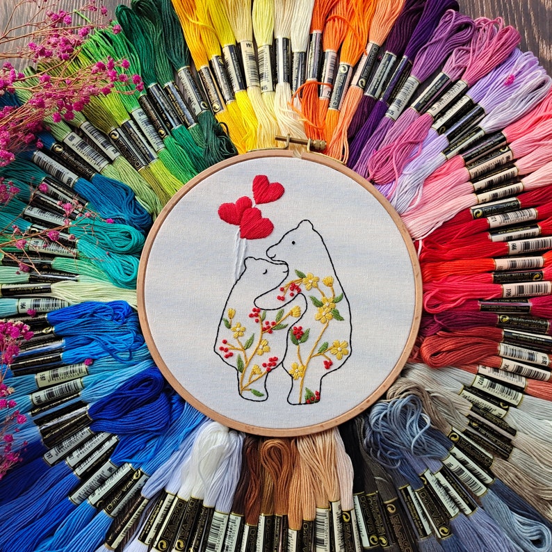 Pu&ograve; includere: Un cerchio da ricamo bianco con un disegno di due orsi che tengono cuori. Gli orsi sono circondati da fiori e foglie. Il cerchio &egrave; circondato da una variet&agrave; di fili da ricamo colorati.