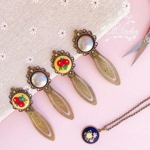 Puede incluir: Cinco marcapáginas dorados con diseños florales bordados. Cada marcapáginas presenta un marco redondo adornado con un diseño floral diferente. Los marcapáginas están dispuestos sobre una superficie rosa.