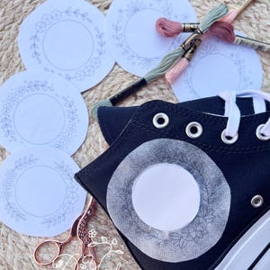 Könnte beinhalten: Ein schwarzer High-Top-Sneaker mit weißen Schnürsenkeln und einer weißen Sohle, mit einem floralen Stickdesign. Mehrere kreisförmige Stickmuster, Stickgarn in Rosa und Grün sowie roségoldene Scheren sind ebenfalls vorhanden.