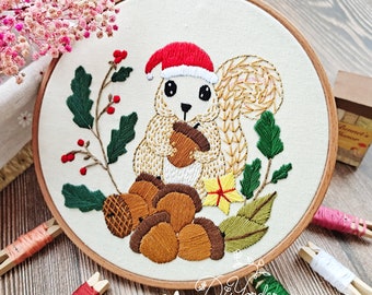 Könnte beinhalten: Stickbild mit Stickerei, das ein süßes Eichhörnchen zeigt, das einen roten Weihnachtsmann-Hut trägt und Eicheln hält. Das Eichhörnchen ist von grünen Blättern und einer einzelnen gelben Blume umgeben. Der Rahmen ist aus Holz und hat einen weißen Stoffhintergrund.