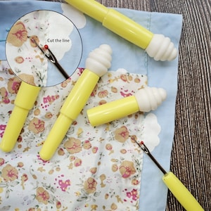Puede incluir: Cuatro enhebradores amarillos con tapas de plástico blanco. Los enhebradores se utilizan para ayudar a enhebrar una aguja. La imagen muestra un primer plano de los enhebradores sobre una tela floral. El texto "Cut the line" es visible en la imagen.