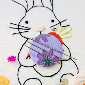 May include: Embroidery needles rest on a purple floral fabric circle, part of an embroidered bunny design. The bunny is outlined in black thread on white fabric, with a carrot and other floral elements. A small pink flower is also present.