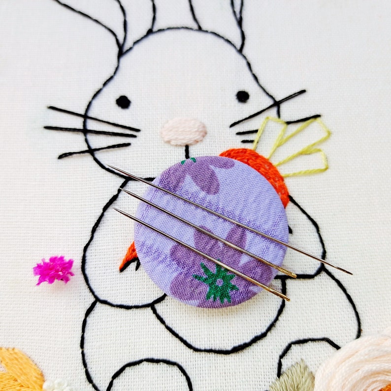 May include: Embroidery needles rest on a purple floral fabric circle, part of an embroidered bunny design. The bunny is outlined in black thread on white fabric, with a carrot and other floral elements. A small pink flower is also present.