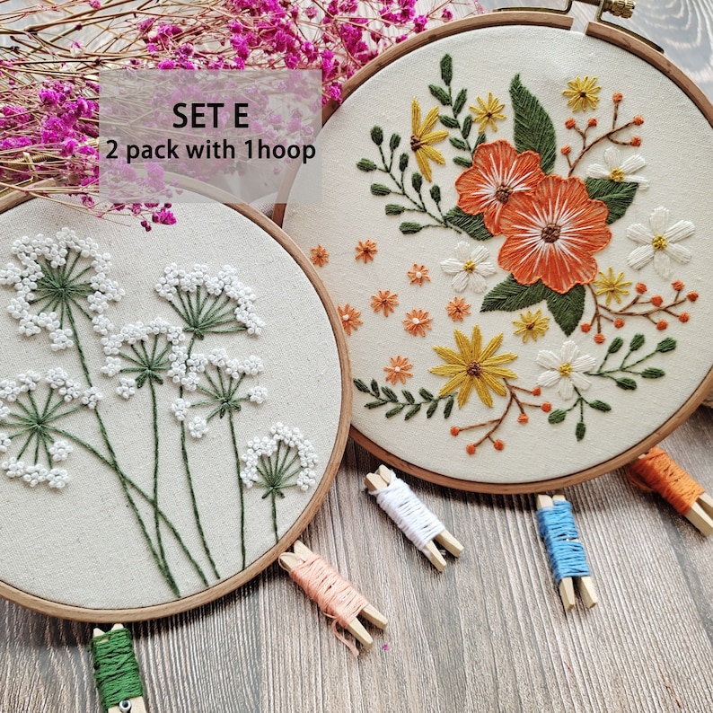 Peut inclure: Deux cerceaux &agrave; broder avec des motifs floraux. Le cerceau de gauche pr&eacute;sente des fleurs blanches avec des tiges vertes. Le cerceau de droite pr&eacute;sente des fleurs orange, jaunes et blanches avec des tiges vertes. Le texte "SET E 2 pack with 1 hoop" est visible sur l'image.