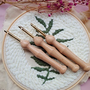 May include: Three wooden yarn needles with gold tips, lying on a white and green embroidery hoop. The needles are used for punch needle embroidery.