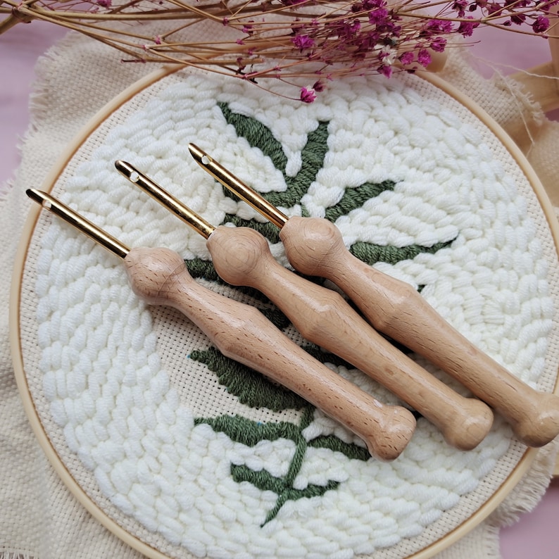 May include: Three wooden yarn needles with gold tips, lying on a white and green embroidery hoop. The needles are used for punch needle embroidery.