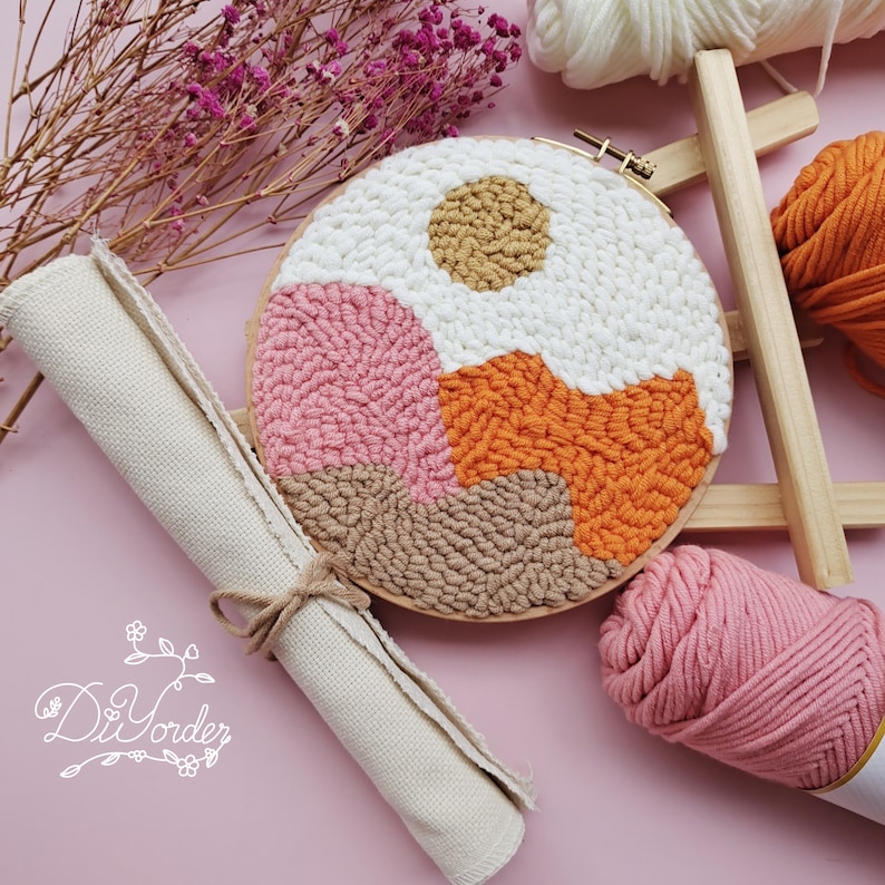 May include: A punch needle kit with a wooden hoop, pink, orange, brown, and white yarn, and a piece of white canvas. The hoop is filled with a punch needle design of a sunset over a landscape.