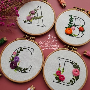 May include: Four embroidery hoops featuring the letters A, B, C, and D embroidered in black thread. Each letter is encircled by a wreath of green leaves and colourful flowers. The flowers are pink, purple, orange, and red.