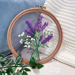 May include: A close-up of a purple and white floral embroidery on a mesh fabric stretched in a wooden embroidery hoop. The embroidery is of lavender flowers and small white flowers.