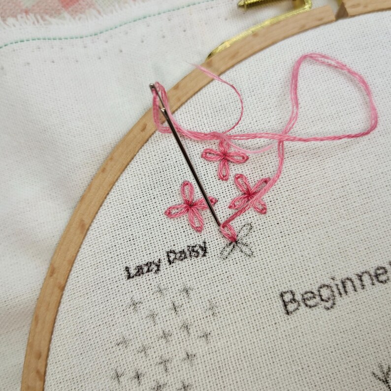 K&ouml;nnte beinhalten: Nahaufnahme eines rosa Stickprojekts in einem h&ouml;lzernen Stickrahmen. Das Projekt ist ein einfaches Design aus rosa Lazy-Daisy-Blumen mit dem Text "Lazy Daisy" und "Beginner" auf dem Stoff.