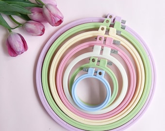 May include: A set of six pastel-colored embroidery hoops, ranging in size from small to large. The hoops are made of plastic and have a smooth finish. They are arranged in a rainbow pattern, with the smallest hoop being light blue and the largest hoop being lavender.