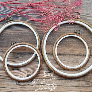 以下が含まれることがあります： 茶色の木製の外枠と白い内枠を持つ、3つの異なるサイズの刺繍枠。枠はピンクの花が咲いた木製の表面の上に置かれています。