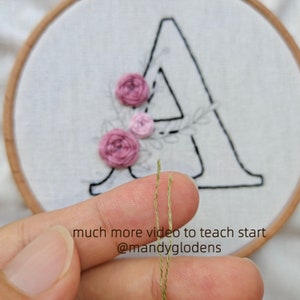 May include: Close-up of two strands of green embroidery floss held between two fingers. The floss is being used to embroider a floral design on a white fabric with the letter "A" embroidered in black. The text "much more video to teach start @mandyglodens" is visible below the floss.