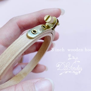 Puede incluir: Un aro de bordado de madera de 7,6 cm de diámetro con un mecanismo de apriete de tornillo de latón. El aro se sostiene en una mano.