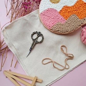 May include: A beige embroidery canvas with a pair of small gold scissors, a ball of yarn, and a completed embroidery hoop with a colorful abstract design.