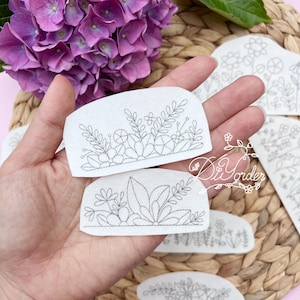 Op de afbeelding: Twee witte stoffen met zwarte lijntekeningen van bloemmotieven. De ontwerpen zijn verschillende arrangementen van bloemen, bladeren en stengels.