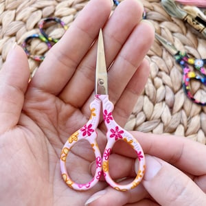 May include: Small, pink and yellow floral embroidery scissors with silver blades. The scissors have a decorative floral pattern on the handles. Other embroidery tools are in the background.