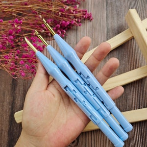 May include: Three blue plastic crochet hooks with gold metal tips. The hooks are held in a hand.