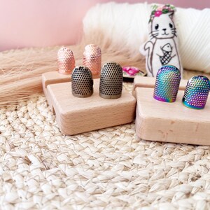 May include: A collection of thimbles in various colors and finishes, including copper, bronze, and iridescent rainbow. The thimbles are displayed on small wooden blocks, with a white yarn ball and a cat illustration in the background.